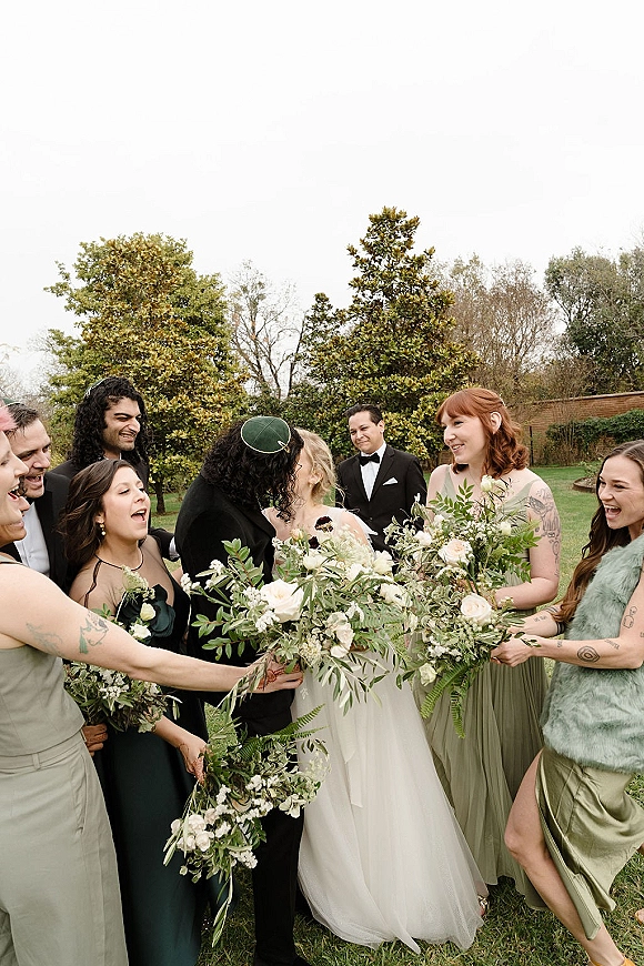 Wedding party portrait of the bride and groom kissing as bridesmaids and groomsmen cheer, with white rose bouquets on a lawn by trees and a brick wall