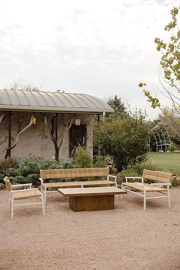 Outdoor lounge seating with wicker bench and armchairs around a wooden coffee table on a gravel patio beside a stone building under clouds