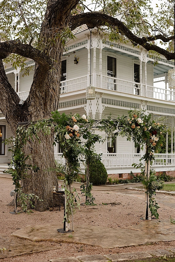 Wedding ceremony arch with greenery garland and blush-peach roses on a wooden frame, set before a Victorian house and large tree lawn