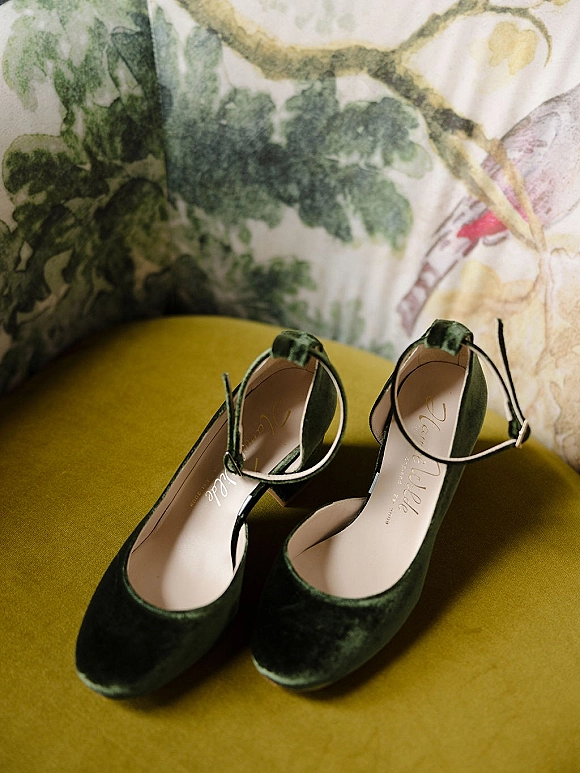 Bridal shoes in green velvet bridal heels with ankle straps resting on a yellow upholstered chair against floral wallpaper backdrop