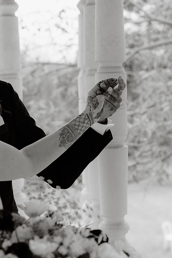 Wedding ceremony vows as henna-adorned hands exchange rings, groom’s tuxedo jacket and boutonniere near gazebo columns and trees