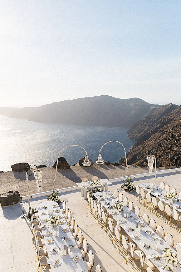 Outdoor reception setup with long white-linen banquet tables, gold rim chairs, florals and lanterns on a cliffside terrace overlooking the ocean