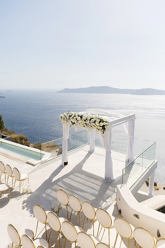 Ceremony setup for an outdoor wedding ceremony with a white floral arch and gold chairs on a cliffside terrace overlooking the ocean