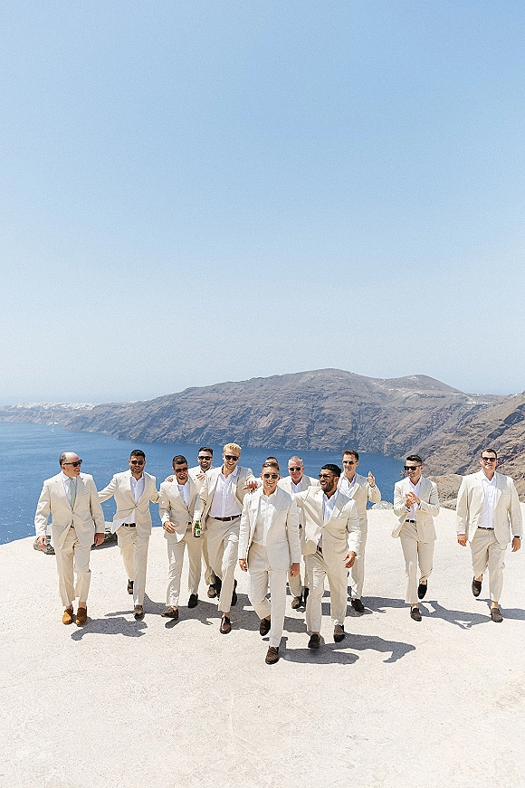 Groomsmen portrait of the group walking in light beige suits and sunglasses on a white stone terrace above the ocean coastline