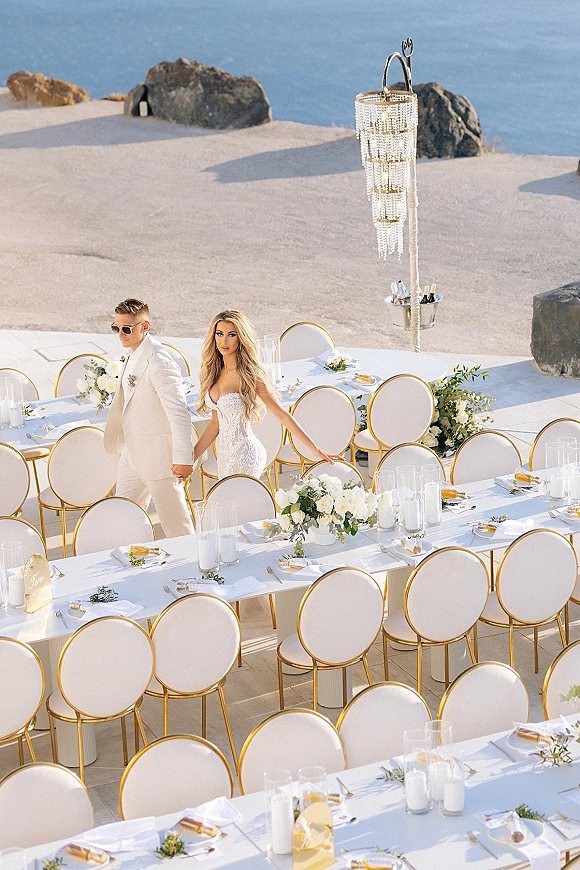 Reception tablescape with long banquet tables, white floral centerpieces and greenery garlands, set on a stone terrace overlooking ocean water