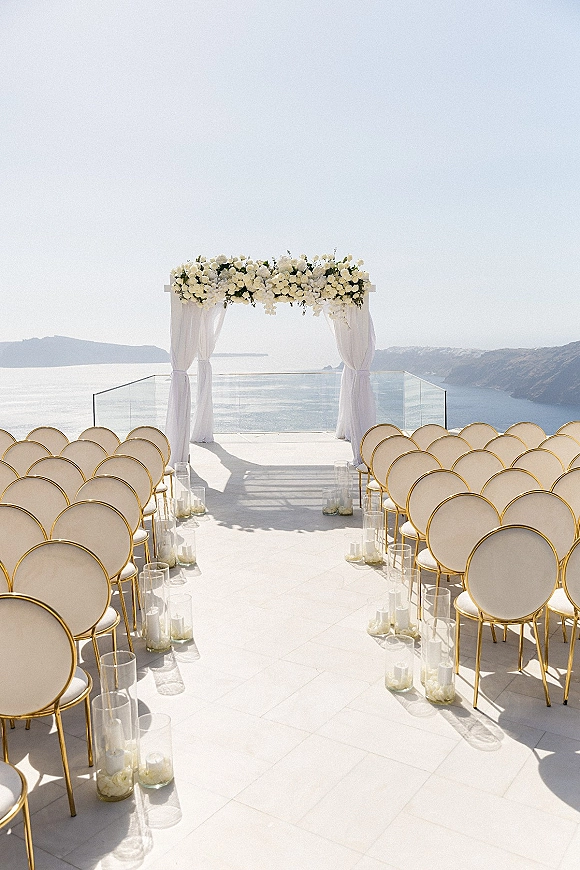 Ceremony setup with a white floral arch and drapery, candle-lined aisle and gold chairs on a terrace overlooking ocean cliffs