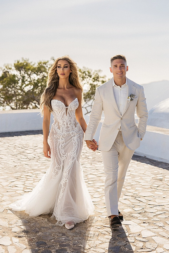 Couple portrait of bride and groom walking hand in hand, her strapless lace mermaid gown with train, his white suit, in a courtyard