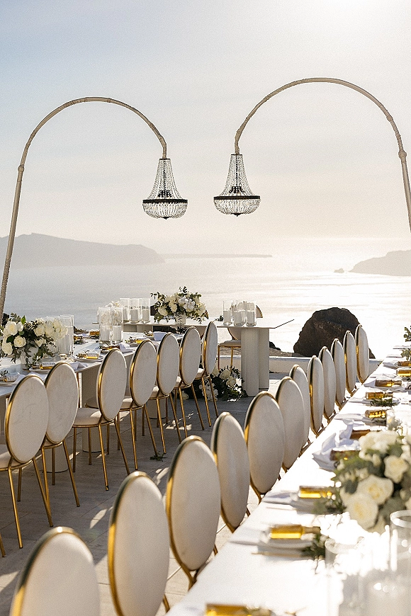Reception tablescape for an outdoor wedding reception with long banquet tables, white florals and greenery under chandeliers on a cliffside terrace overlooking the ocean