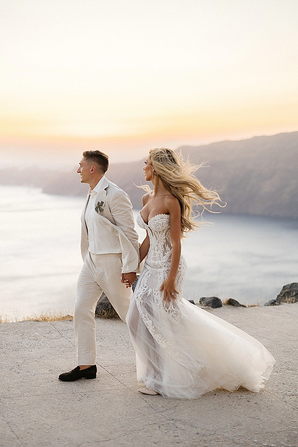 Couple portrait holding hands on a cliffside wedding portrait with ocean view at sunset, bride in strapless lace dress and groom in suit