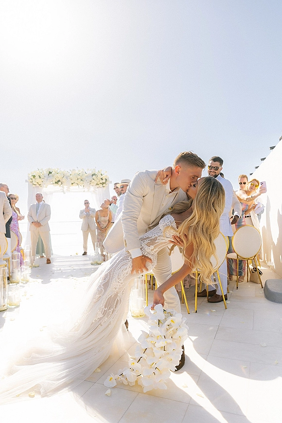 Wedding kiss portrait of bride and groom in a dip kiss beneath a white floral arch on an oceanfront terrace, guests watching under blue sky