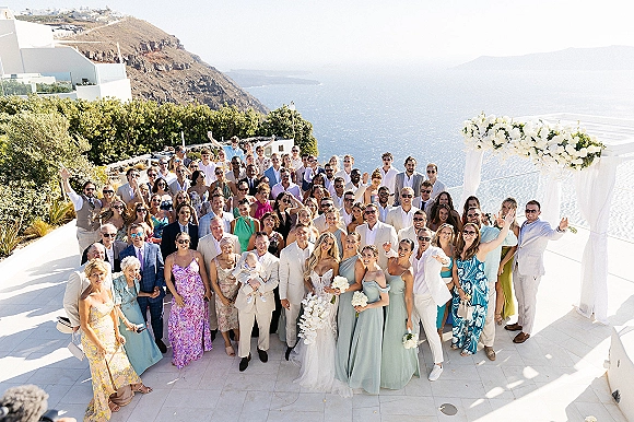Wedding group photo of bride and groom with guests under a white floral arch, ocean-view cliffside terrace behind, many in sunglasses