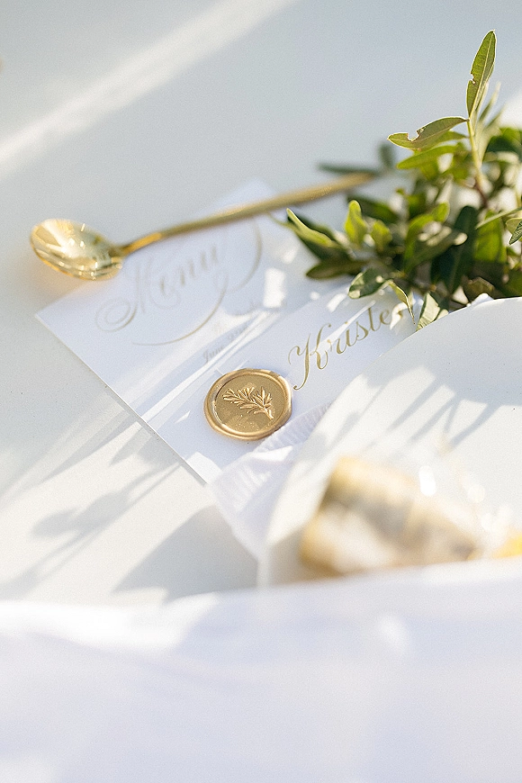 Invitation suite wedding invitation flat lay with vellum wrap, gold calligraphy, wax seal, greenery, and gold spoon on white in sun shadows