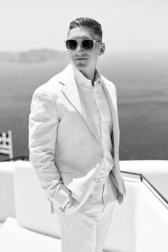 Groom portrait in a white suit wearing sunglasses, leaning by the yacht railing with ocean and wide sky in the background