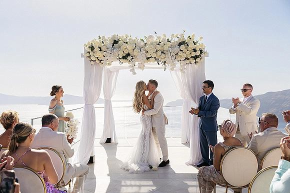 Wedding kiss under a white floral arch with draped fabric on a terrace, ocean and mountains behind, guests in sunglasses applauding
