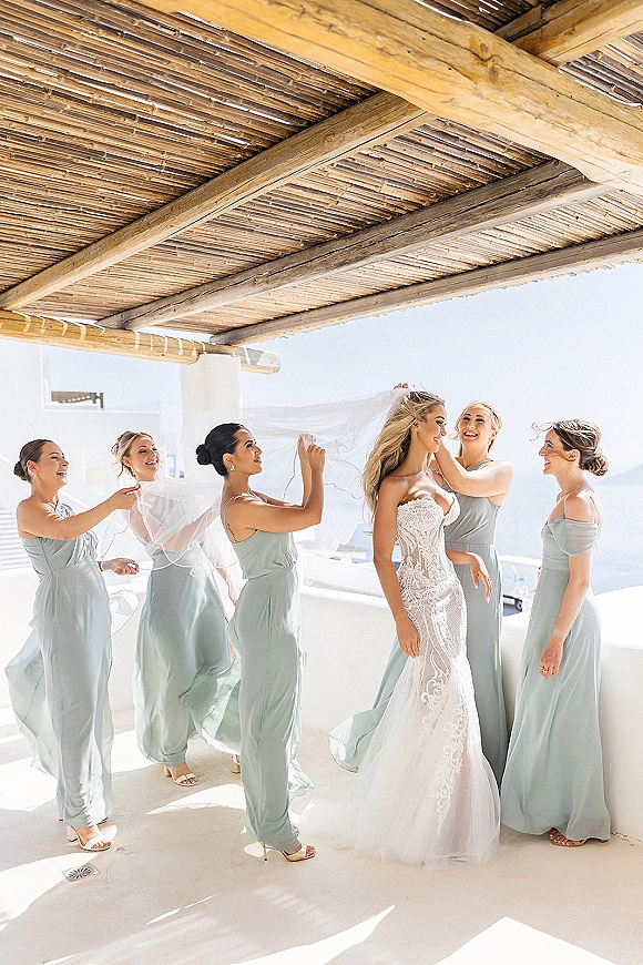 Bride getting ready as bridesmaids adjust her bridal veil, lace gown flowing on an ocean-view terrace with white stucco walls and pergola