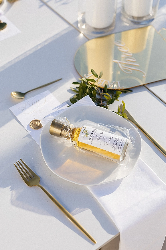 Wedding place setting with gold flatware, white plate and napkin, olive sprig, acrylic menu and wax-sealed escort card in sunlit shadows