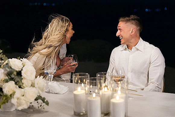 Reception toast moment as the bride and groom laugh during a wedding speech reaction at an outdoor night table under bistro string lights, candlelit decor