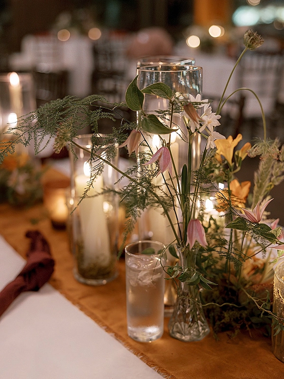 Reception tablescape with wedding candle centerpiece of glass cylinder vases, pillar and votive candles, greenery and wildflowers under warm bistro lights