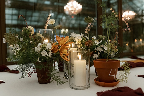 Reception centerpiece with wildflower wedding centerpiece in terracotta pots, orange lilies and pillar candles on a table under string lights and chandeliers