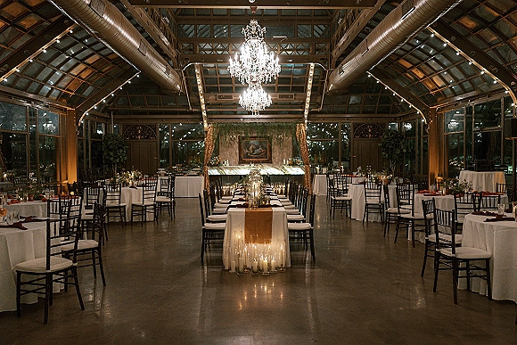Reception tablescape with a long banquet table wedding setup, candles in glass cylinder vases and floral centerpieces under chandeliers in a greenhouse space