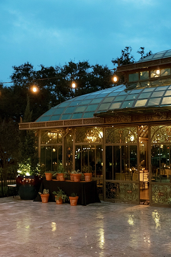 Glasshouse reception venue with glass conservatory wedding venue doors, string lights, and potted plants on a patio at dusk under trees