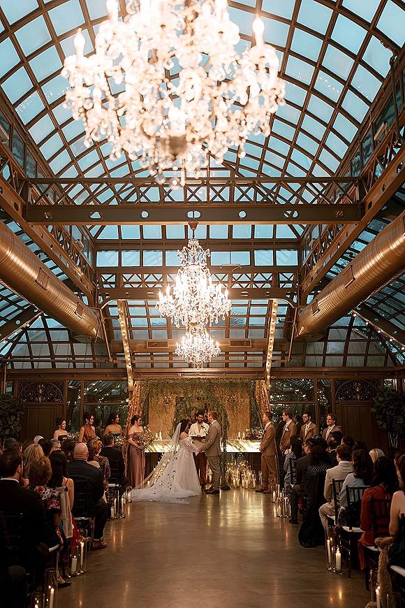 Wedding ceremony under crystal chandeliers with aisle candles, bride and groom at a greenery arch in a glass-ceiling conservatory with guests seated