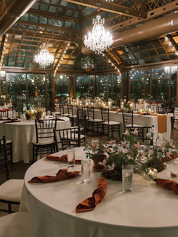 Reception tablescape in a greenhouse wedding reception with white linens, rust napkins, candlelit centerpieces, chandeliers and string lights at night