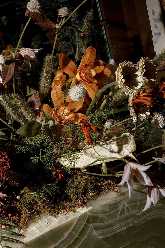 Wedding floral arrangement with orange orchid centerpiece, daisies, dried seed pods and moss on a stone surface in a moody dark interior