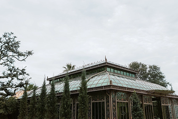 Wedding venue exterior with a glass conservatory featuring ornate ironwork trim and large windows, set among trees under a cloudy sky