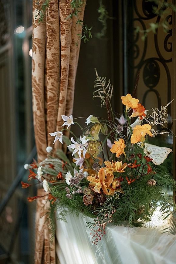 Wedding flower arrangement with orange and white blooms, greenery, and red berries on a pedestal with draped fabric by window and curtains