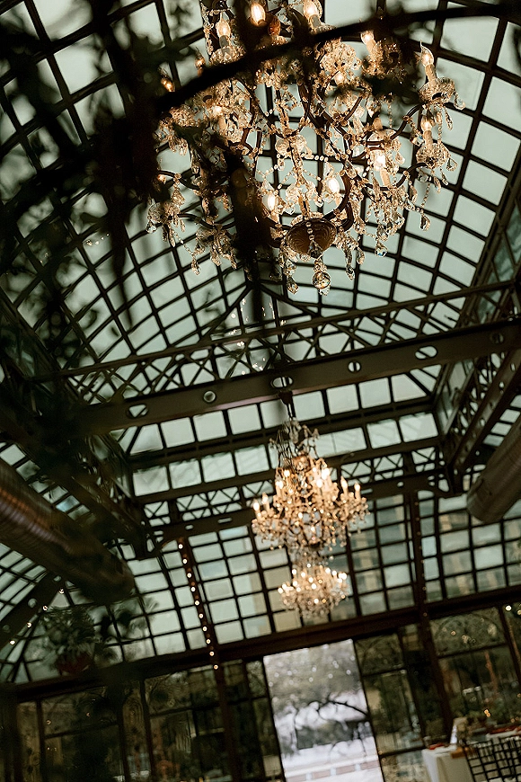 Wedding venue ceiling with crystal chandeliers under a glass ceiling, metal grid beams, and string lights among greenhouse greenery