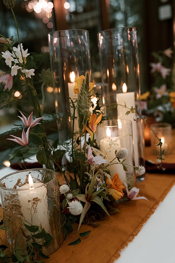 Reception centerpiece with candle centerpiece wedding pillar candles in glass hurricane vases, wildflowers and greenery on a rust runner under chandeliers by windows