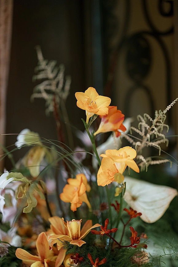 Wedding floral arrangement with orange orchid centerpiece and lush greenery, fern fronds, and grasses against a dark interior backdrop