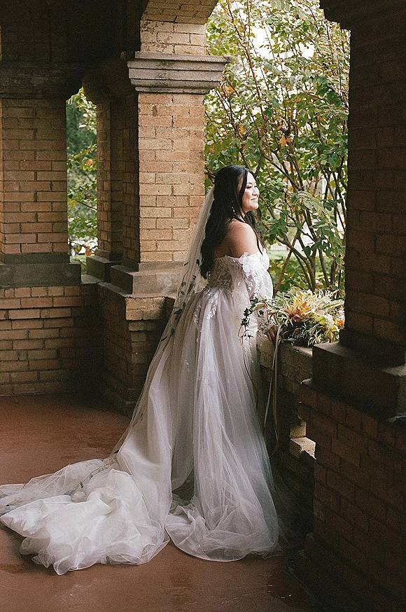 Bridal portrait of a bride holding bouquet with trailing ribbons, in side profile with cathedral veil and long train under brick arches