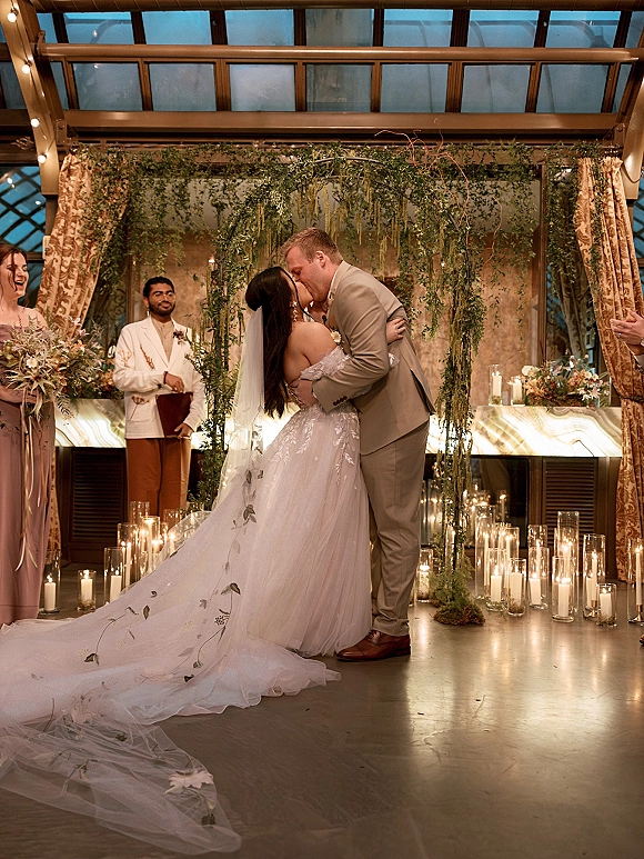 Wedding kiss as the couple embraces at the altar under a floral arch, with veil and candles in glass cylinders beneath a glass ceiling