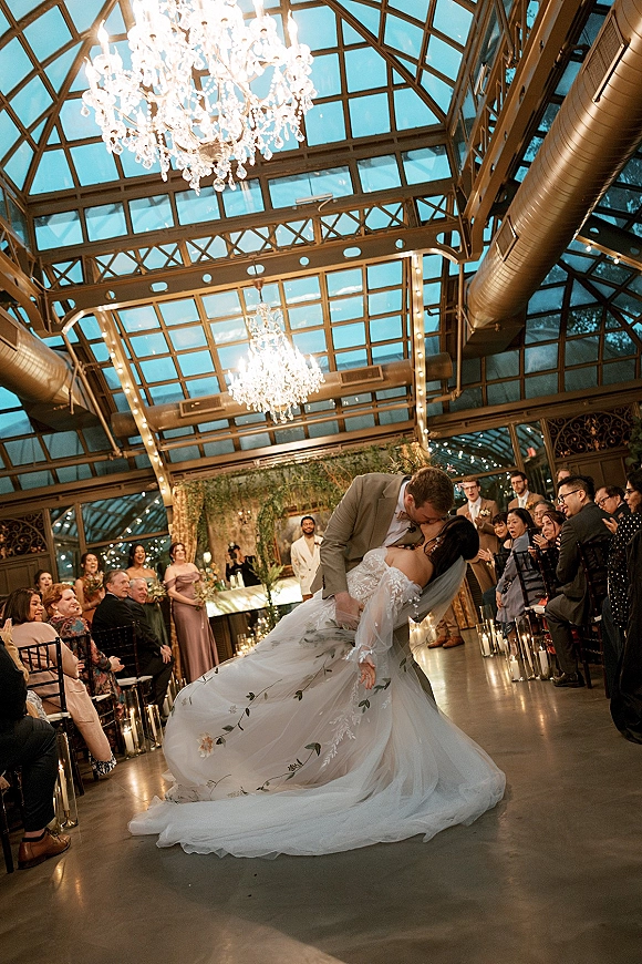 Wedding kiss as groom dips the bride in veil and dress amid aisle candles, under chandeliers and string lights in a glass conservatory