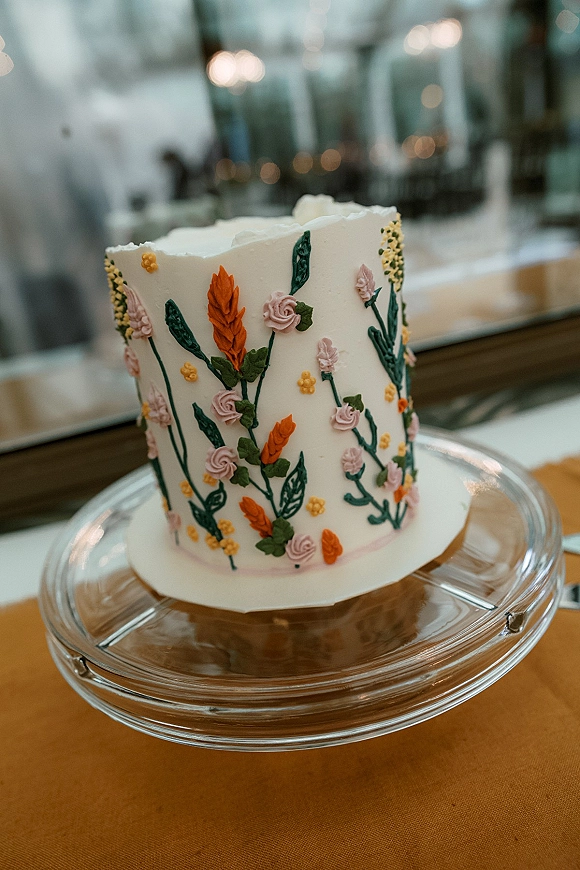 Wedding cake with buttercream wedding cake frosting and piped floral icing on a glass stand, set indoors by a mirror under bistro lights