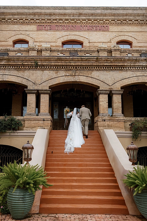 Wedding recessional as bride and groom walk up stairs from behind, her long veil trailing as he leads toward a brick arched entryway