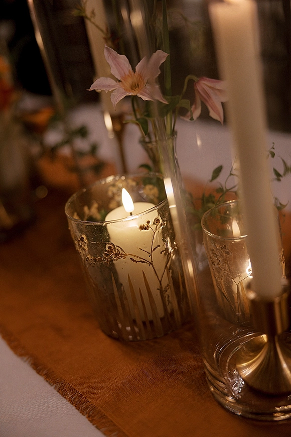 Wedding table centerpiece with taper candles and a pink flower in a bud vase on a linen runner, glowing on a warmly lit reception table