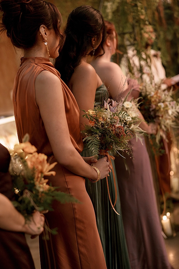 Bridesmaid lineup holding bouquets in mismatched bridesmaid dresses with ribbon ties, standing by aisle candles in a garden ceremony