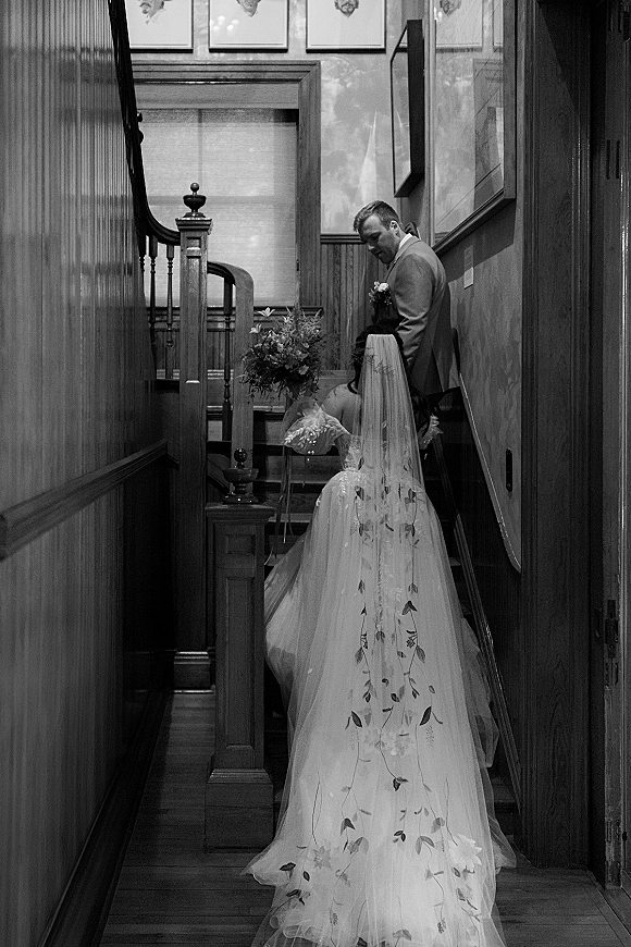 First look moment as groom waits on stairs while bride approaches in wedding dress and long embroidered veil, bouquet in hand, wood-paneled hallway