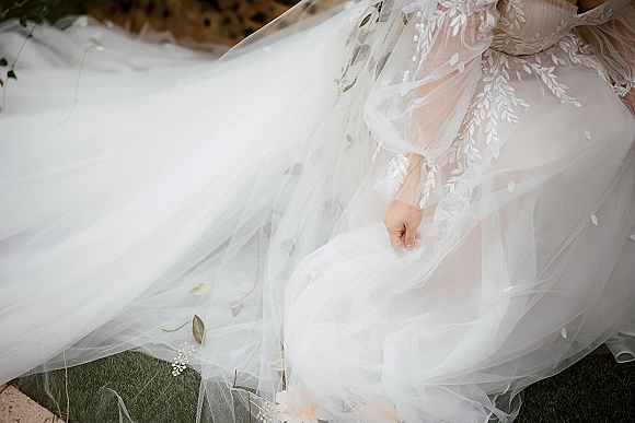 Wedding dress close-up showing lace wedding dress details, tulle skirt and floral appliqué with illusion sleeves, on grass near a wicker chair