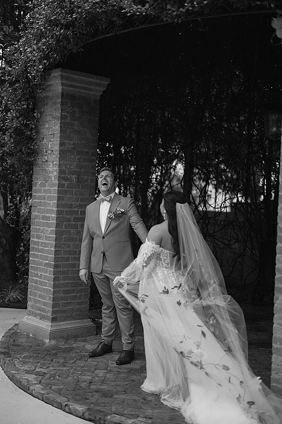 First look moment as bride in lace wedding dress and long veil taps groom in bow tie by a wrought iron gate in brick courtyard garden
