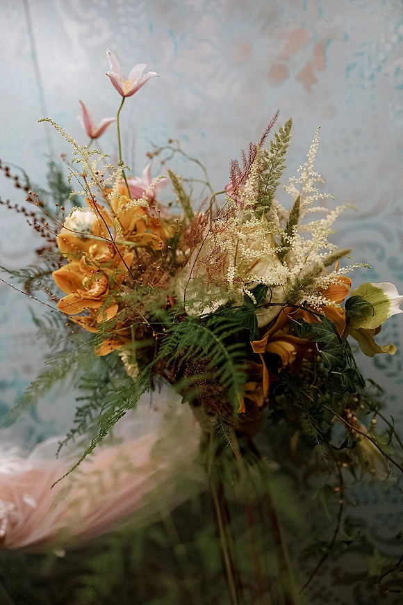 Bridal bouquet with orange orchids and pink tulips, white astilbe, and ferns, trailing greenery against patterned wallpaper indoors