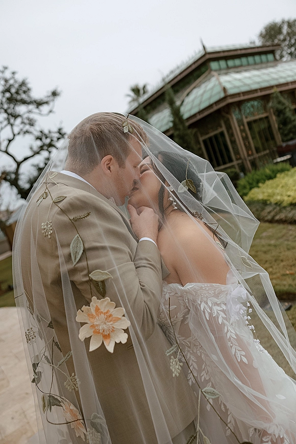 Wedding kiss portrait of bride and groom in a veil kiss photo, floral embroidered veil over off-shoulder dress on garden lawn by greenhouse
