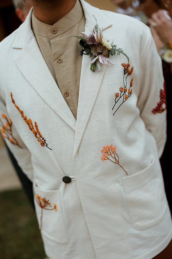 Groom suit detail with embroidered wedding suit lapel, floral embroidery and boutonniere, beige shirt and buttons, guests blurred behind