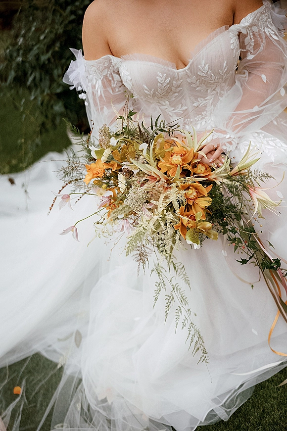 Bridal bouquet with orange wedding bouquet blooms, white wildflowers, and trailing greenery held against an off-shoulder embroidered gown on grass