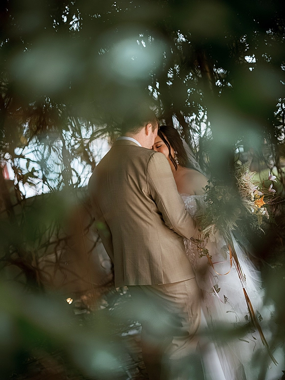 Wedding couple portrait of bride and groom embrace, her veil and wildflower bouquet against soft outdoor greenery and tree branches