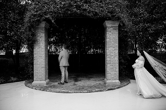Wedding first look as bride approaches groom from behind, long veil and off-the-shoulder lace dress in brick courtyard garden walkway
