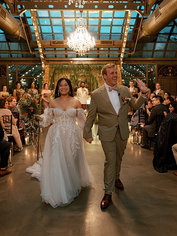 Wedding recessional as bride and groom walking aisle holding hands, groom waving under glass ceiling with chandelier and candlelit aisle chairs